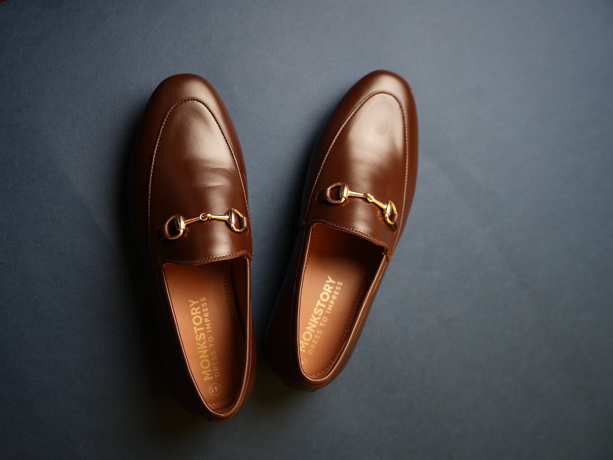 A pair of brown leather loafers with gold-tone horsebit detailing displayed on a dark background.