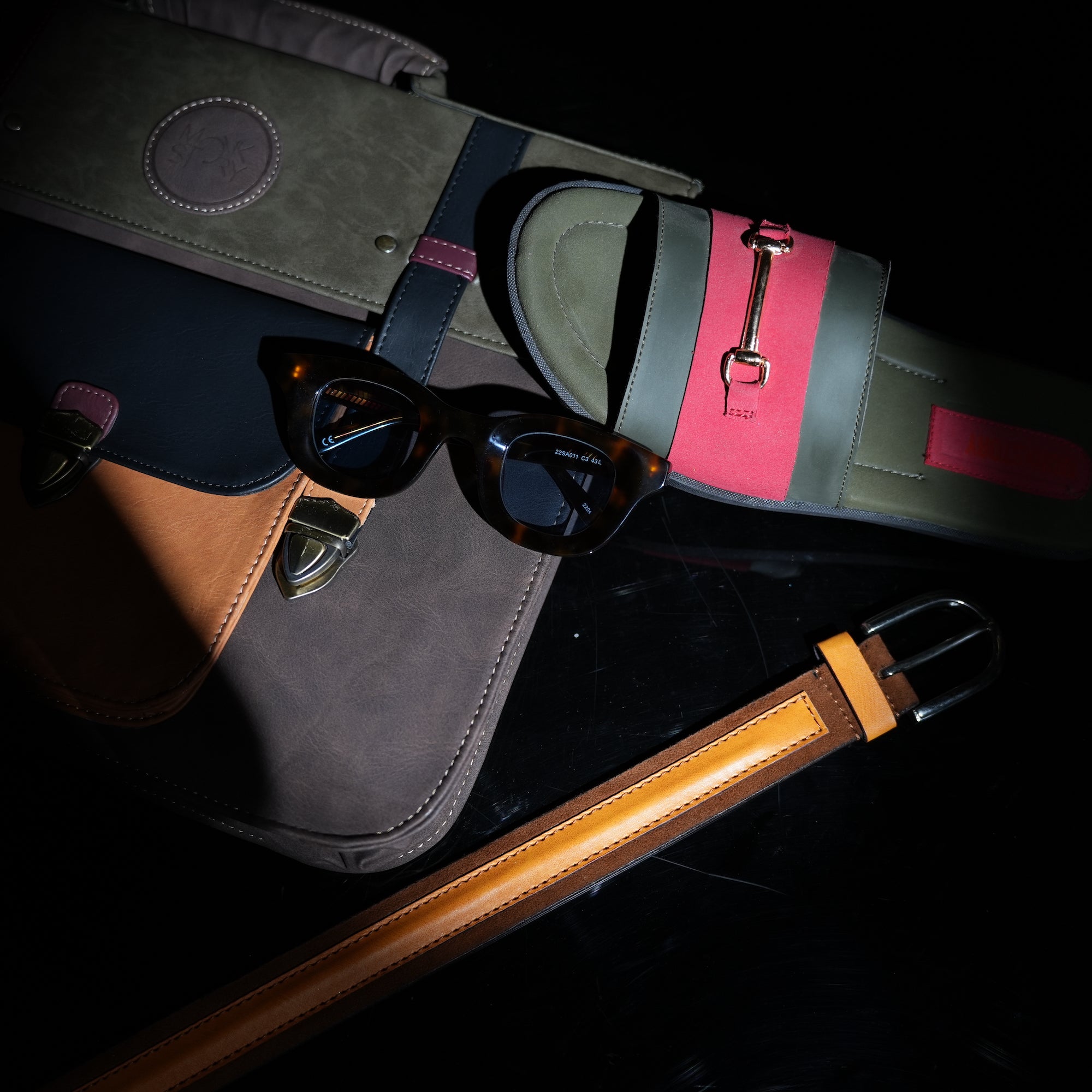 A bag, sunglasses, and a belt are laying on a dark surface.