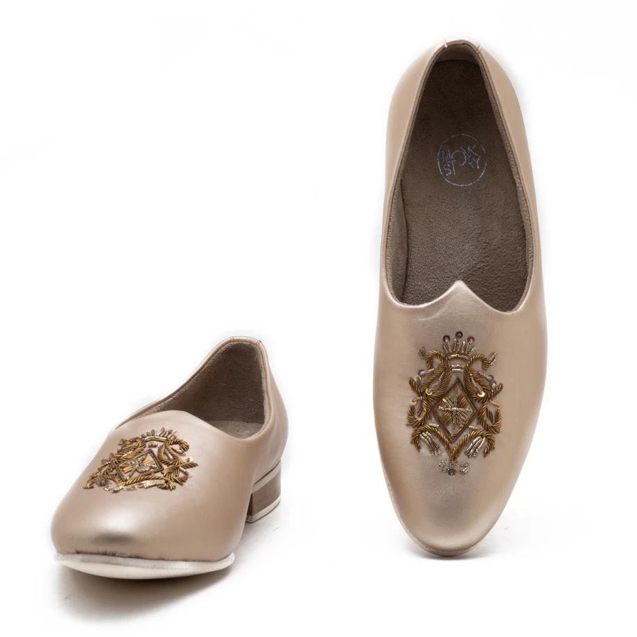 A pair of Monkstory Magenta Mojri - Cream shoes with a gold embroidered crest.